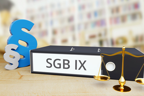 Dekorativ: Auf dem Bild ist eine schwarze Aktenmappe mit der Aufschrift „SGB IX“ zu sehen, die auf einem hellen Holztisch liegt. Rechts neben der Aktenmappe steht eine goldene Waage, die Symbol für Gerechtigkeit und Rechtsprechung ist. Links von der Aktenmappe befinden sich zwei Paragraphensymbole, eines in Blau und eines in Grau. Im Hintergrund ist ein unscharfes Bücherregal zu erkennen.