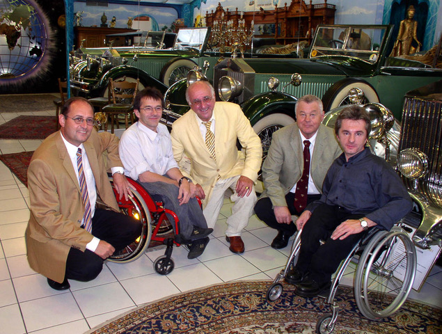 Fünf Männer posieren in einem Ausstellungsraum mit klassischen Oldtimern. Zwei der Männer sitzen in Rollstühlen, die anderen drei knien oder hocken neben ihnen. Im Hintergrund sind mehrere grüne, glänzende Oldtimer zu sehen sowie dekorative Wandmalereien und Teppiche auf dem Boden. Die Stimmung wirkt freundlich und respektvoll.