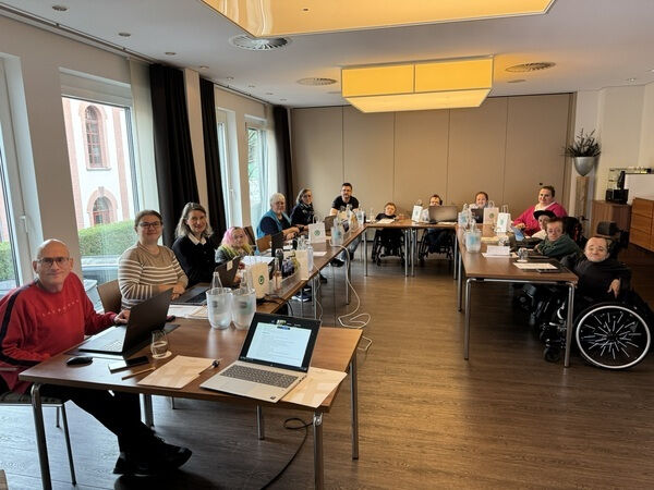 Eine Gruppe von etwa zwölf Menschen sitzt in einem hellen Konferenzraum an mehreren zusammengestellten Tischen. Einige Personen arbeiten an Laptops, andere lächeln in die Kamera. Unter den Teilnehmenden sind auch Personen im Rollstuhl. Auf den Tischen stehen Getränke, Unterlagen und technische Geräte. Durch große Fenster fällt Tageslicht in den Raum.