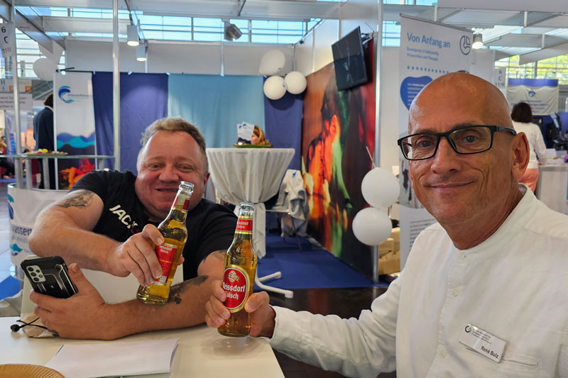 Das Bild zeigt zwei Männer am Messestand der Deutschen OI-Gesellschaft (DOIG) auf der Rehacare-Messe 2023. Links sitzt Martin Schmidt, ein lächelnder Mann mit kurz geschnittenem, hellem Haar und einem schwarzen T-Shirt. Er hält eine Flasche Reissdorf Kölsch in der Hand und hat ein Smartphone in der anderen Hand. Rechts neben ihm sitzt René Bulz, der eine Brille und ein weißes Hemd trägt und ebenfalls eine Flasche Reissdorf Kölsch hält. Beide stoßen fröhlich an und lächeln in die Kamera.