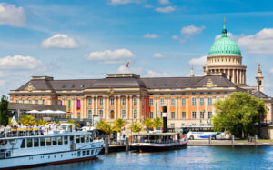 Blick auf das Potsdamer Stadtschloss mit der großen grünen Kuppel der St.-Nikolaikirche im Hintergrund. Davor liegen Ausflugsschiffe am Ufer der Havel, unter blauem Himmel mit weißen Wolken.